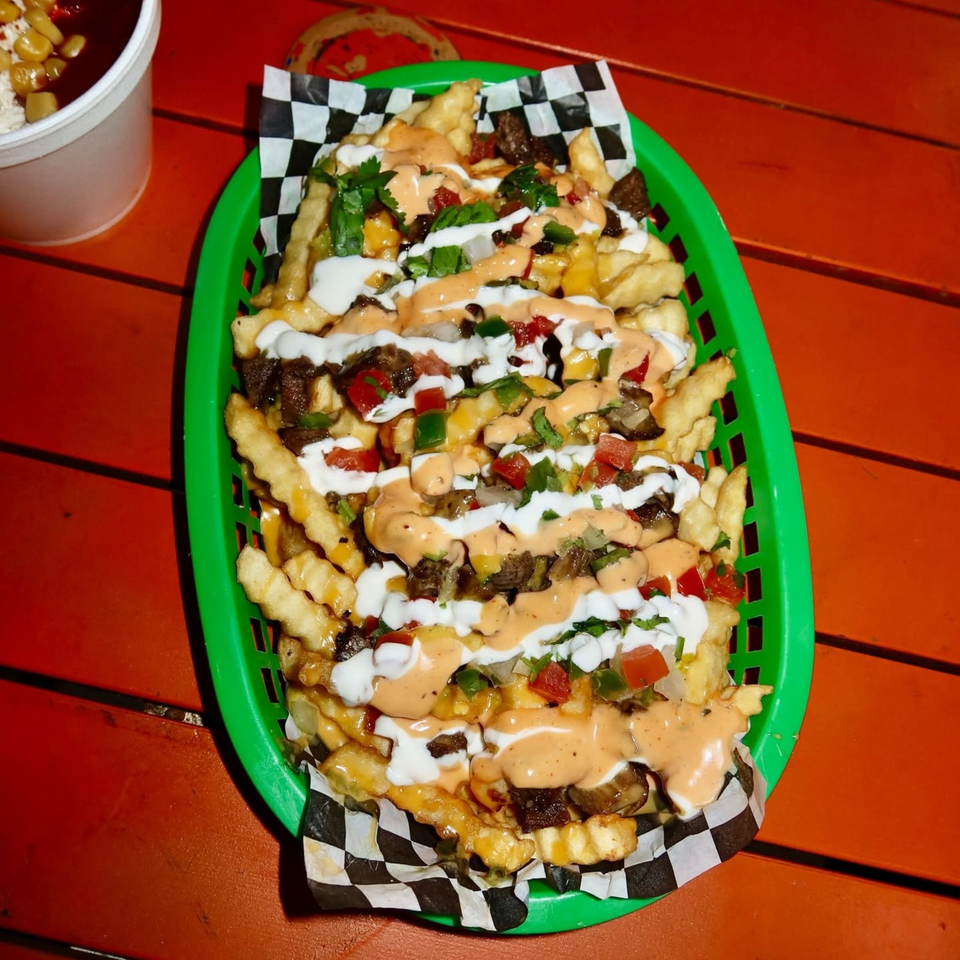 Loaded crinkle-cut fries topped with grilled meat, cheese, tomatoes, peppers, herbs, and creamy white and orange sauces in a green basket.