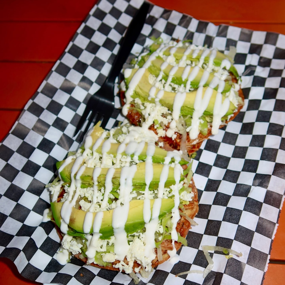 Two loaded tacos topped with avocado, crumbled cheese, lettuce, and white crema on checkered paper.