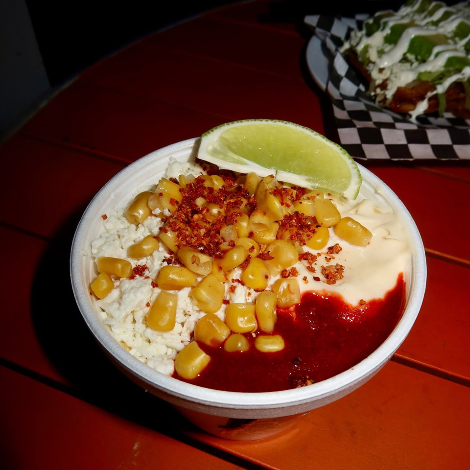 Bowl of creamy dip topped with corn, crumbled bacon, red sauce, and lime wedge on a red table.