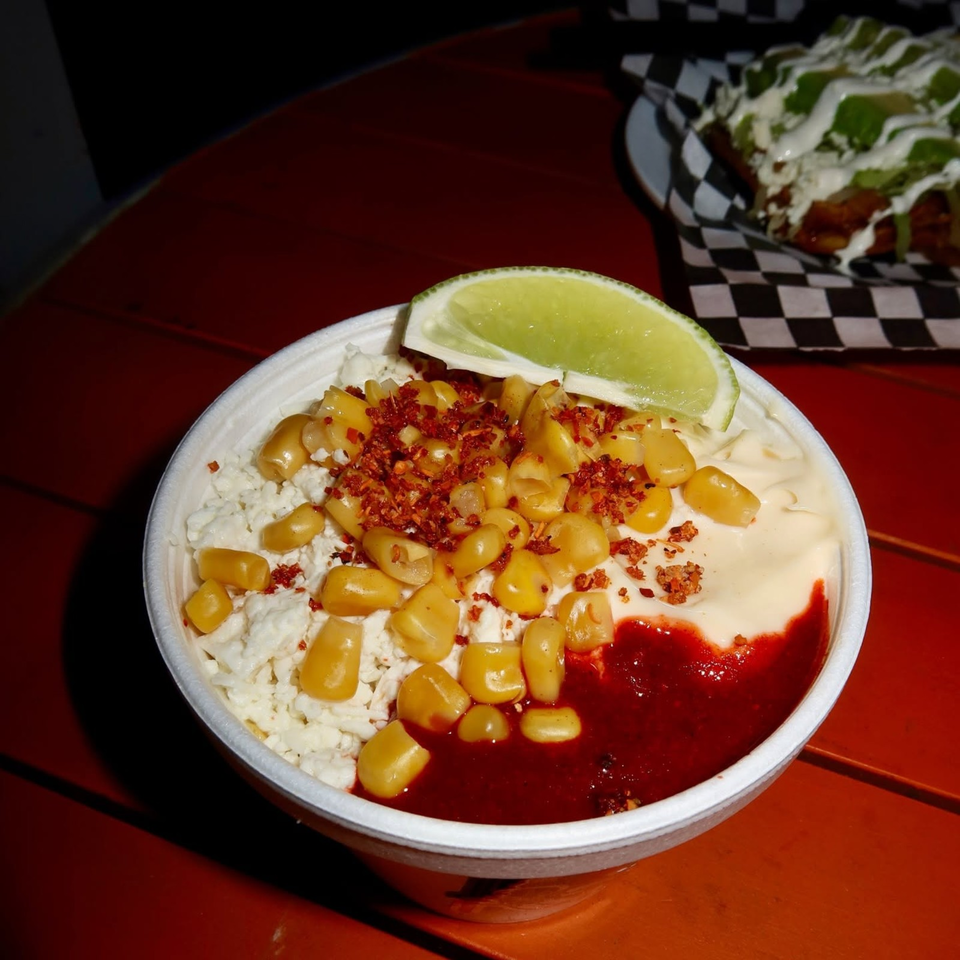Bowl of creamy dip topped with corn, crumbled cheese, chili flakes, red sauce, and a lime wedge on a wooden table.