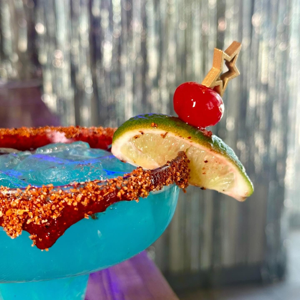Colorful cocktail garnished with a lime wedge, red cherry, and chili-coated rim in a blue glass.